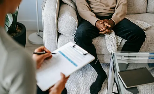 CBT psychotherapy session with therapist holding clipboard and patient sitting on couch, hands clasped, discussing mental health treatment