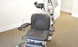 Medical examination chair with armrests, headrest, footrest, and attached equipment, used for TMS therapy in an eye clinic or similar medical setting