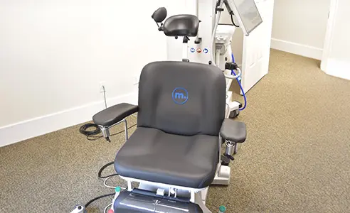 Medical examination chair with armrests, headrest, footrest, and attached equipment, used for TMS therapy in an eye clinic or similar medical setting