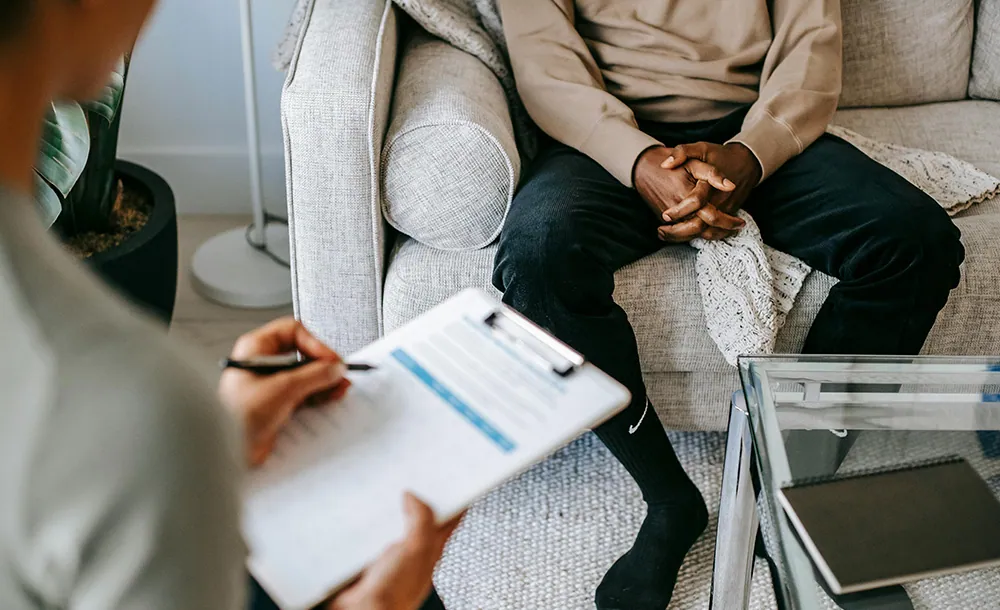 CBT psychotherapy session with therapist holding clipboard and patient sitting on couch, hands clasped, discussing mental health treatment