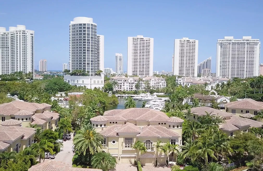 Aerial view of luxury homes, palm trees, and high-rise condominiums in Aventura, Florida, with a marina and boats in the background on a clear sunny day.