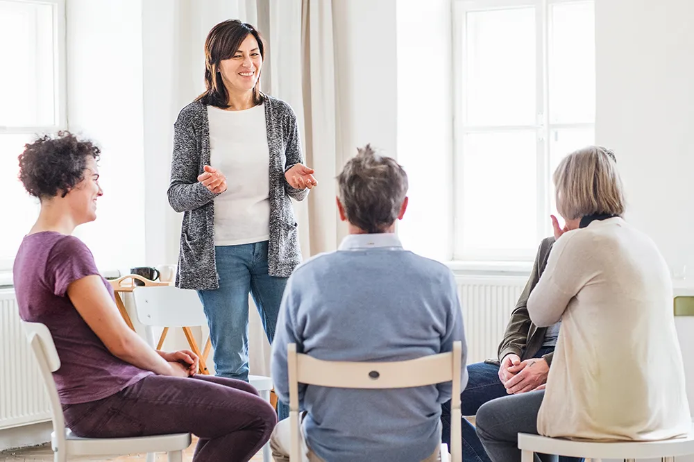 A group of people are participating in a group psychotherapy session. They are seated in a circle, engaging in open and supportive discussions led by a therapist. The setting is calm and welcoming, emphasizing the collaborative and healing nature of the therapy.