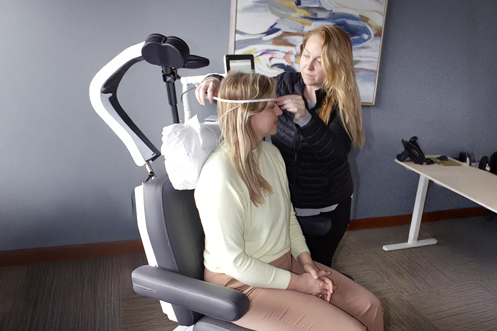 A healthcare professional is administering Transcranial Magnetic Stimulation (TMS) therapy to a patient. The patient is seated comfortably while the professional adjusts the TMS device, which is positioned near the patient's head. The setting is calm and clinical, emphasizing the therapeutic nature of the treatment.