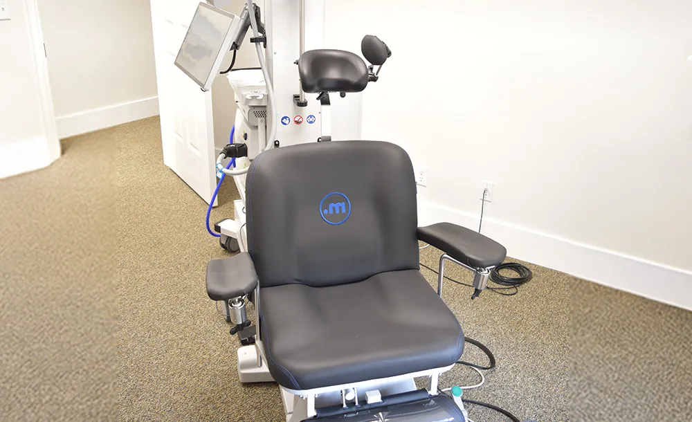 Medical examination chair with armrests, headrest, footrest, and attached equipment, used for TMS therapy in an eye clinic or similar medical setting