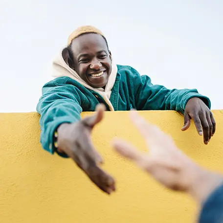 A smiling man with light colored hair reaches out his hand over a bright yellow wall towards another person, symbolizing support and connection during Men's Mental Health Awareness Month.
