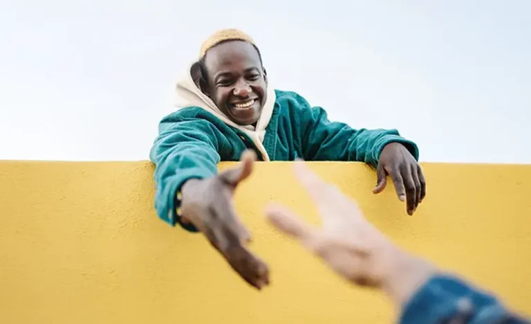 A smiling man with light colored hair reaches out his hand over a bright yellow wall towards another person, symbolizing support and connection during Men's Mental Health Awareness Month.