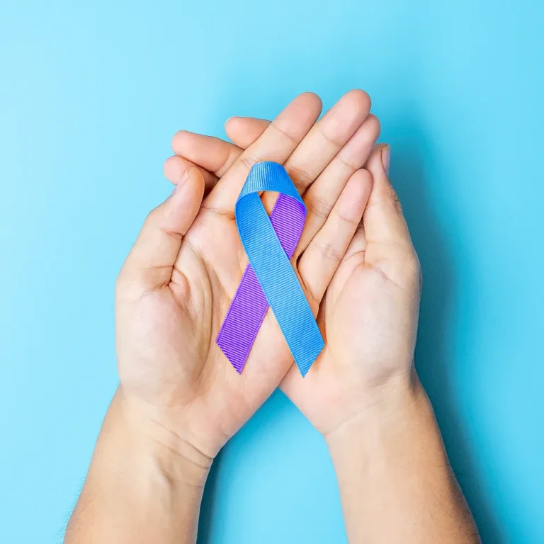 A pair of hands gently cup a ribbon against a light blue background. The ribbon is half purple and half blue, symbolizing suicide prevention and awareness.