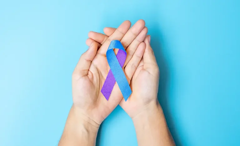 A pair of hands gently cup a ribbon against a light blue background. The ribbon is half purple and half blue, symbolizing suicide prevention and awareness.
