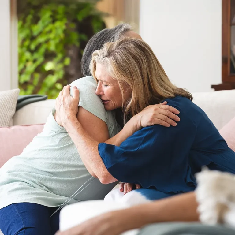 Two older women embrace in a deep, comforting hug on a sofa, symbolizing mutual emotional support and finding help during a time of grief or loss.