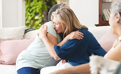 Two older women embrace in a deep, comforting hug on a sofa, symbolizing mutual emotional support and finding help during a time of grief or loss.