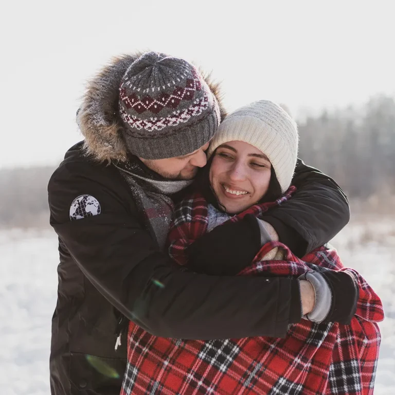 A happy couple embraces closely outdoors in the bright winter snow, both wearing warm knit hats and scarves. The man is hugging the smiling woman from behind.